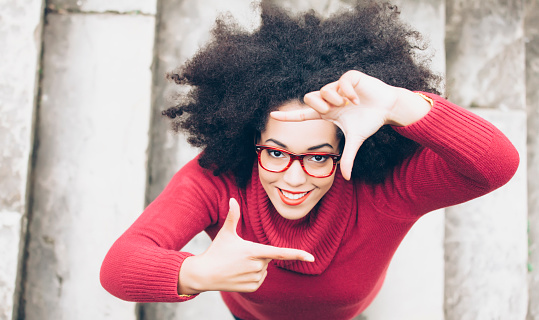 Portrait of smiling young woman standing on staircase. Looking at camera and making finger frame. High angle view. Woman with black curly hair, red eyeglasses, and red pullover.