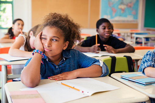 Teenage girls and boy sitting in a row at the desks in the classroom and writing an exam. Focus on pensive girl, looking away with hand on chin.