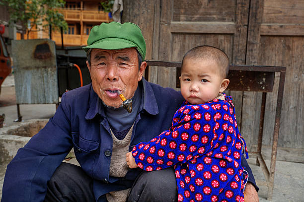 Little boy hugging his grandfather on rural street, Guizhou, China. Zhaoxing Dong Village, Guizhou Province, China - April 9, 2010: Three year child hugging an elderly man who holds in his mouth pipe smoking. dong stock pictures, royalty-free photos & images