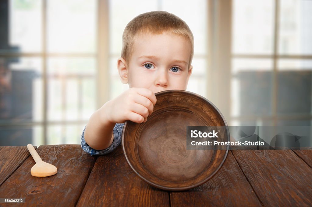 Cute blonde boy shows empty plate, hunger concept Child hunger concept. Small toddler boy shows empty bowl sitting at dark wood table with wooden spoon. Cute child has no food in plate Child Stock Photo