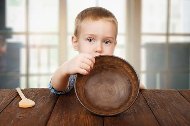 Child hunger concept. Small toddler boy shows empty bowl sitting at dark wood table with wooden spoon. Cute child has no food in plate