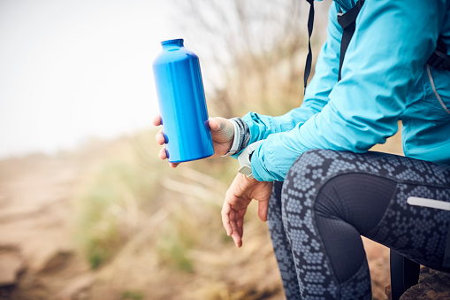 Midsection of traveler holding water bottle. Female is wearing casuals. She is sitting on rock.