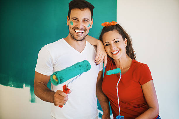 Couple decorating apartment. Closeup front view portrait of a happy early 30's couple standing in front of partially painted wall, looking at camera and smiling. Both holding paint rollers. The guy has some paint on his face. mural photos stock pictures, royalty-free photos & images