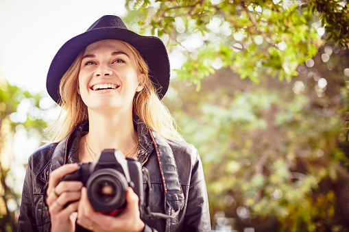 Smiling young woman holding SLR camera. Blond female hipster is wearing hat while looking away. She is wearing leather jacket outdoors.