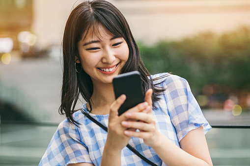 Smiling young Japanese woman with smart phone outdoors at urban environment.