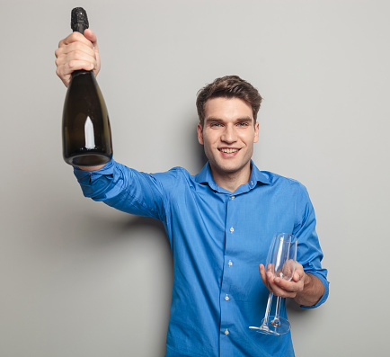 Attractive young man holding a bottle of champagne in his right hand and two glasses in his left hand.