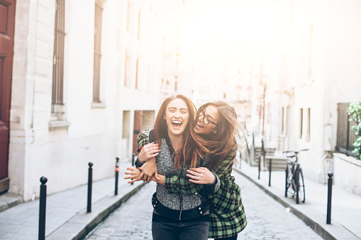 Cheerful young women having fun and embracing on street. Both with long hair, one with coat and eyeglasses. Urban scene and building exterior, a bike and stairs on background.