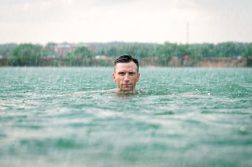 Middle aged man swimming in lake with blue water under the rain in thunderstorm