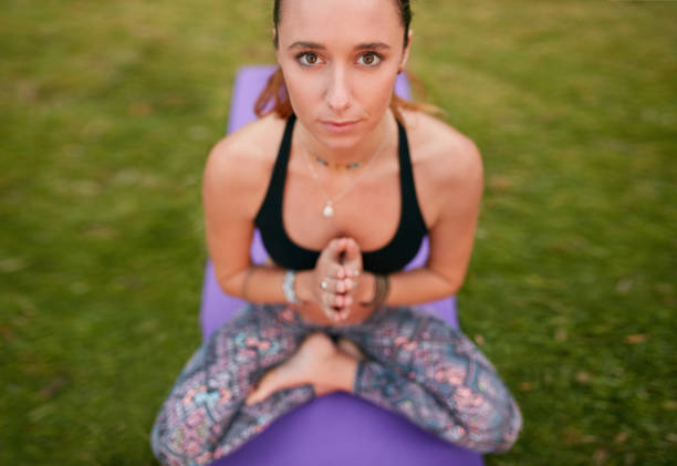 Top view of fitness woman sitting on exercise mat with her hands joined and looking at camera. Fitness woman doing yoga outdoors.