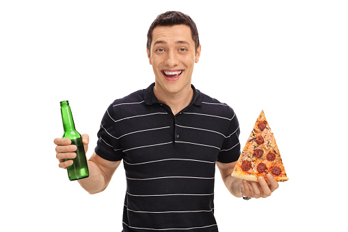 Smiling young guy holding a beer bottle in one hand and a slice of pizza in the other isolated on white background