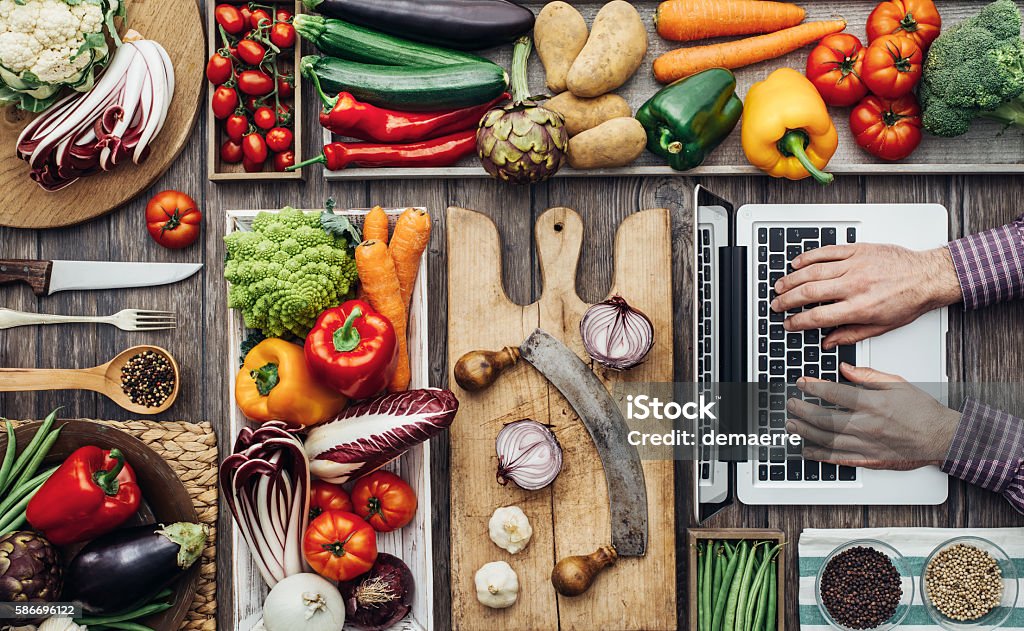 Cooking meets technology Freshly harvested vegetables, cooking utensils and a laptop on a rustic kitchen worktop, a man is searching recipes online Food Stock Photo