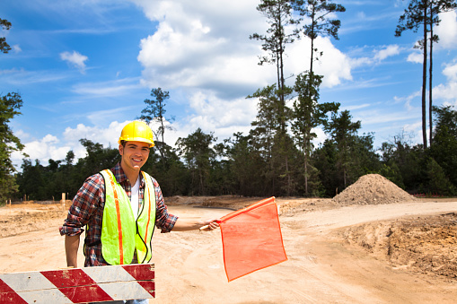 A Latin descent construction worker holds an orange flag to help direct traffic through a construction zone.  Cleared road with barricades in background of newly cleared land site for housing development.  He is wearing a hard hat and safety vest.