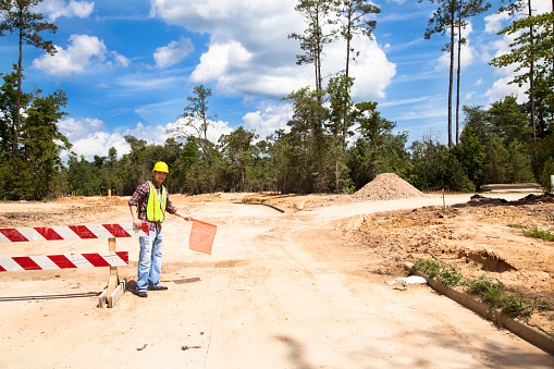 A Latin descent construction worker holds an orange flag to help direct traffic through a construction zone.  Dirt road with barricades in background of newly cleared land site for housing development.  He is wearing a hard hat and safety vest.