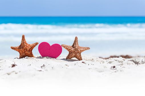 Red sweet heart shape with two starfishes on white sandy beach with sea background. Happy Valentines Day at summertime on exotic vacations.