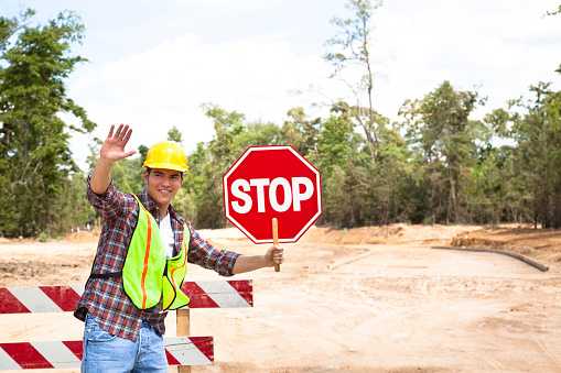 A Latin descent construction worker holds a stop sign to help direct traffic through a construction zone.  Dirt road with barricades in background of newly cleared land site for housing development.  He is wearing a hard hat and safety vest.