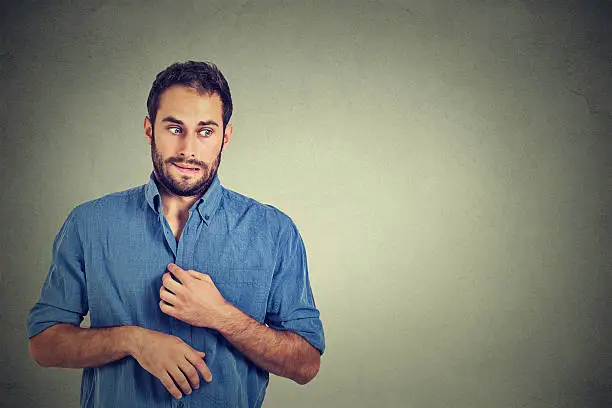 Portrait young man opening shirt to vent, it's hot, unpleasant, awkward situation, playing nervously with hands. Embarrassment. Isolated gray background. Negative emotions facial expression feeling