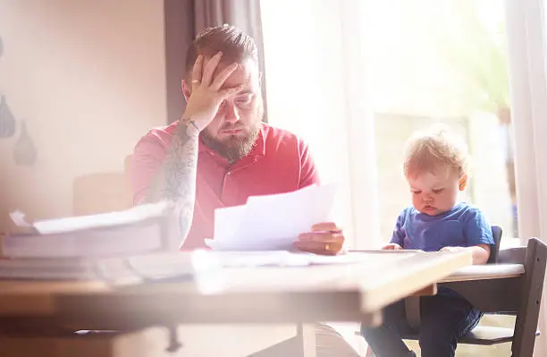 A father sits with his toddler at the dining table. He is reading through his latest bank statements and final demands. He his head in his hands trying to work out how to settle his financial difficulties. His son is sat in his highchair next to him.