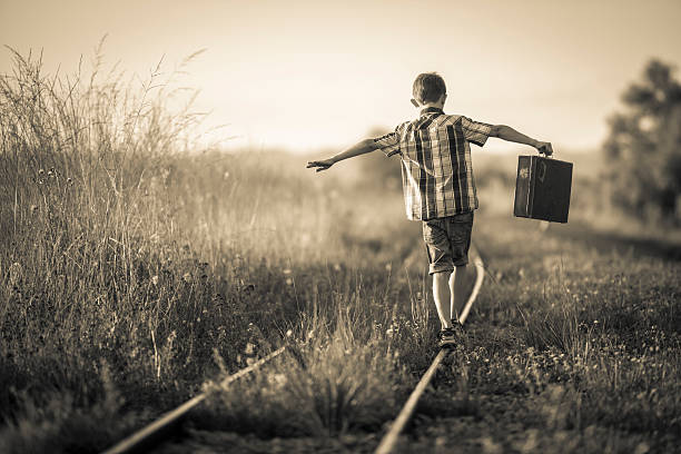 boy with bag balancing on rails Rear view of a boy holding a suitcase in his outstretched arms. His is balancing jauntily on rails in the evening sun. Selective focus on the boy, photo is sepia toned and vignette added for retro style. railroad track photos stock pictures, royalty-free photos & images