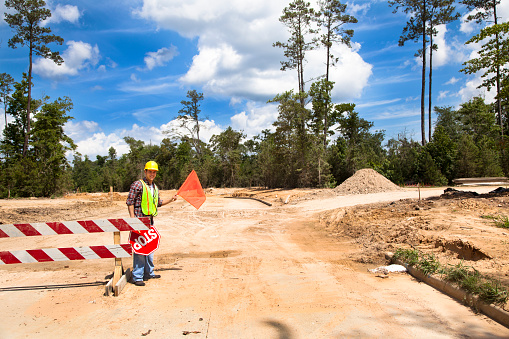 A Latin descent construction worker holds an orange flag and stop sign to help direct traffic through a construction zone.  Dirt road with barricades in background of newly cleared land site for housing development.  He is wearing a hard hat and safety vest.