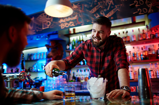 Happy barman looking at client while pouring him whiskey in bar