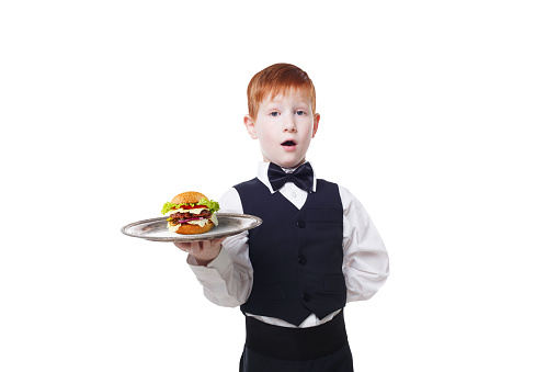 Little surprised waiter stands with tray serving hamburger. Redhead child boy in suit plays restaurant servant, gives burger isolated at white background