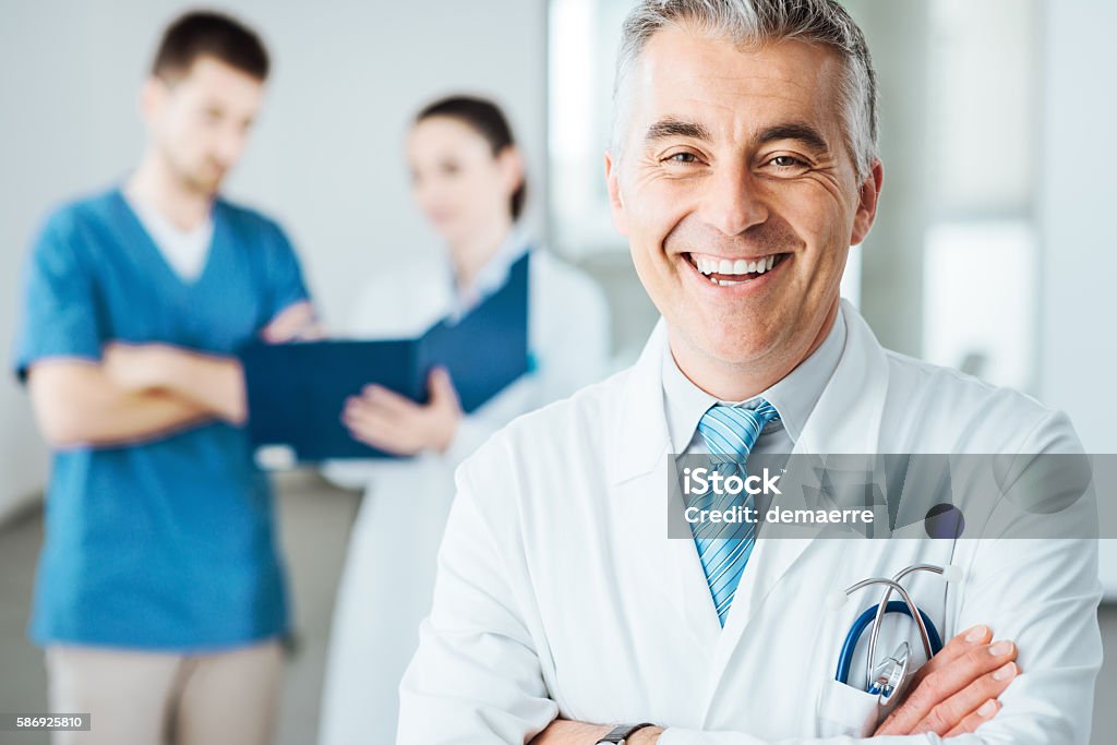 Confident doctor at hospital posing Confident doctor posing and smiling at camera and medical staff checking medical records on background Doctor Stock Photo