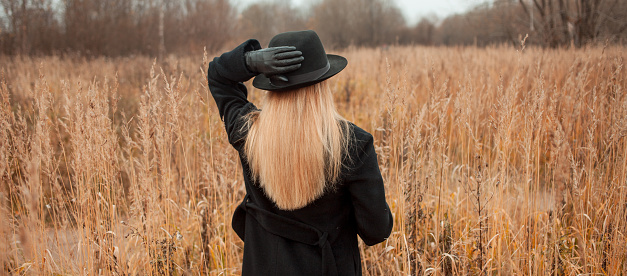 Portrait of young attractive woman in a black coat and hat. She's one in a field reading a book, autumn landscape, dry grass