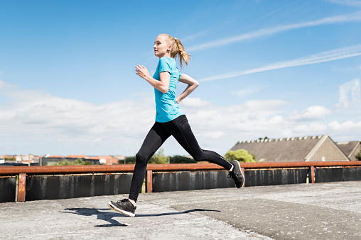 Dynamic portrait of a young woman running in an urban environment. Photographed from a low angle as she passes past she has both feet off the ground, with a shallow focus effect with the point of focus being on her face. She is wearing black tight fitting athletic leggings and a short sleeved pale blue top. She has fair coloured hair pulled back in a pony tail and a fresh clean complexion. Colour, horizontal format with lots of copy space in the blue sky behind her.