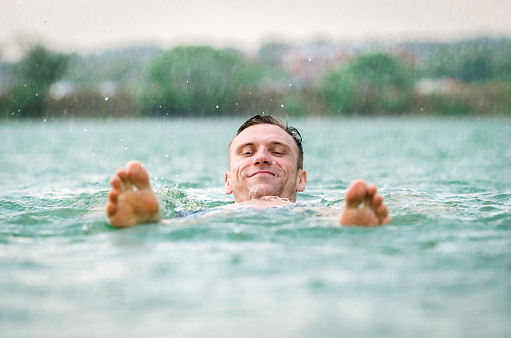 Middle aged man laying on water in blue lake under the rain in thunderstorm.