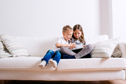 Little girl and boy sitting on sofa with a tablet. Happy children playing indoors.