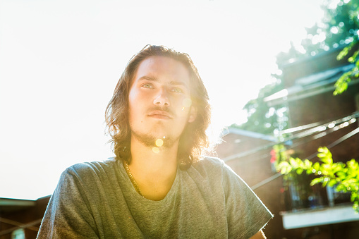 Pensive male teenager on balcony back lit with lens flare. He has long hair and a short beard and moustache.
