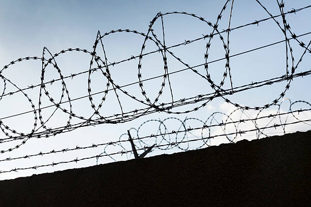 istock Barbed wire fence around prison walls blue sky in background 587186238