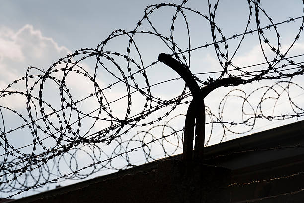 istock Barbed wire fence around prison walls blue sky in background 587186638