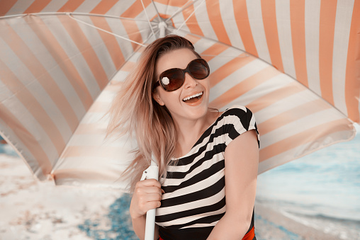 lifestyle shot of young blond hair woman wearing sunglasses and laughing, looking at camera, the photo is taken on the beach, she;s holding red with white umbrella.