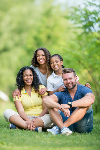 A mixed race family is sitting together in the grass on a sunny summer day. They are smiling and looking at the camera.