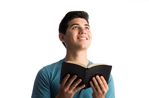 A young latin man holding the Bible open, looking up and smiling in a horizontal waist up shot with white background.