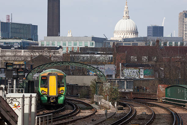 London, England - March 16 2016: Train driver arrives with a Southern Train Service GTR St Paul's Cathedral. Green and yellow passenger train arrives into Elephant and Castle Station in Lindon. Buildings can be seen, St Paul's Cathedral Dome and the tower of the Tate Modern Art Gallery. The four tracks pass over a green bridge. Graffiti tag is on a power box to the bottom left of the image. 