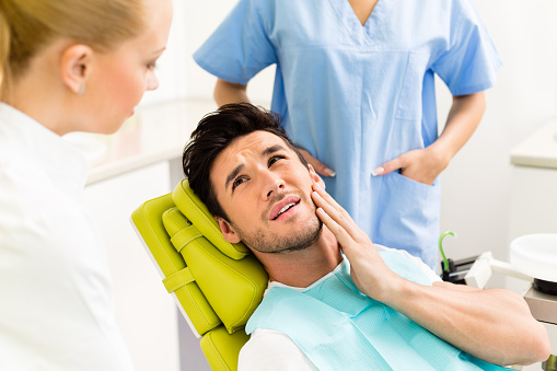 Young man holding cheek in chair at dentist, having toothache.