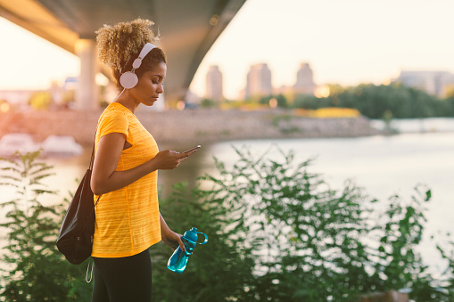 Latina Runner running in the city near river. Walking under the bridge, holding water bottle and typing on her smart phone. Carrying gym bag. Cityscape in background. Wearing big white headphones.