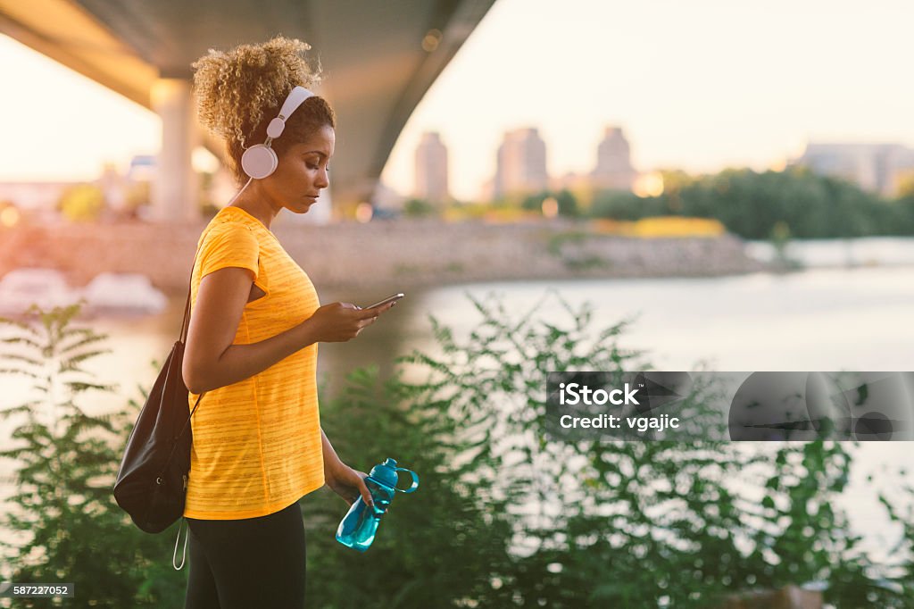 Latina Runner Latina Runner running in the city near river. Walking under the bridge, holding water bottle and typing on her smart phone. Carrying gym bag. Cityscape in background. Wearing big white headphones. Walking Stock Photo