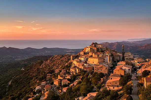 The Balagne village of Speloncato in Corsica bathed in late evening sunshine with the Regino valley and Mediterranean sea behind and pink, orange and deep blue skies above