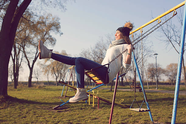 Have fun with yourself! Beautiful caucasian young woman with a hat and scarf swinging in the park playground in autumn sunny day cardigan wales stock pictures, royalty-free photos & images