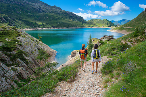 Two friends during a hike in the mountains walking on path near an alpine lake