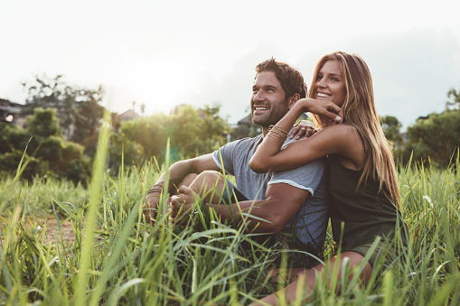 Affectionate young couple sitting on rural grass and looking away smiling. Young man and woman sitting in field looking at view.