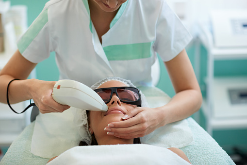 Beautician giving laser epilation treatment to young woman's face at beauty clinic, hairless smooth and soft skin
