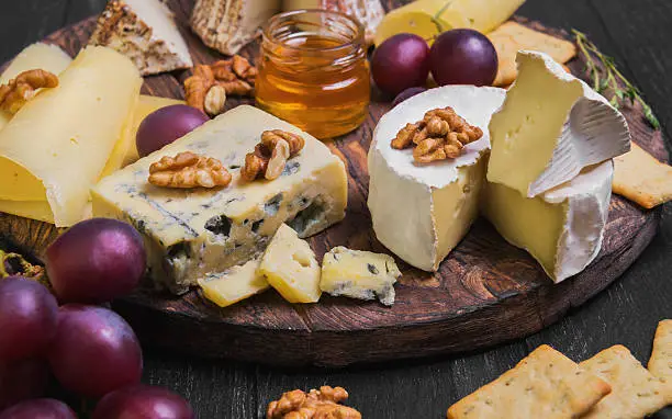 Assorted cheeses on round wooden board plate. Camembert cheese, blue cheese, goat cheese in the grated bark of oak, hard cheese slices, walnuts, grapes, crackers, bread, thyme, honey sauce, dark black wood background