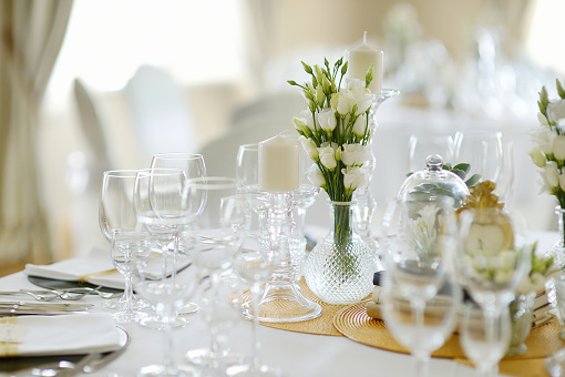 Table arrangement for a special event such a wedding or a celebration with perfectly lined up cutlery and dinnerware with cone-folded cotton napkins, enhanced by floral centerpieces and white candles.