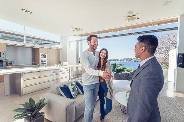 Real estate agent with couple in luxury home. Real estate agent with couple in luxury home. They are shaking hands. There is a water view, kitchen and living room in the background. Couple are casually dressed. They are laughing. Agent is dressed in a suit and smiling. Wide angle. house rental photos stock pictures, royalty-free photos & images
