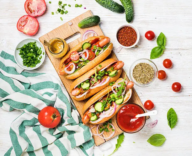 Homemade hot-dogs on wooden serving board with fresh vegetables, spices, ketchup and mustard over white painted old wooden background, top view