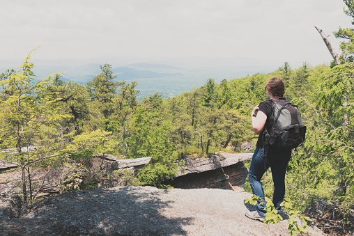 This is a horizontal, color photograph of woman hiking stopping to enjoy the view from Sam's Point Preserve in the Shawangunk Mountains. These mountains are part of the Appalachian Mountains. Green trees fill the valley below the cliffs of the mountain ridge in upstate New York, Ulster County. Photographed with a Nikon D800 DSLR camera.